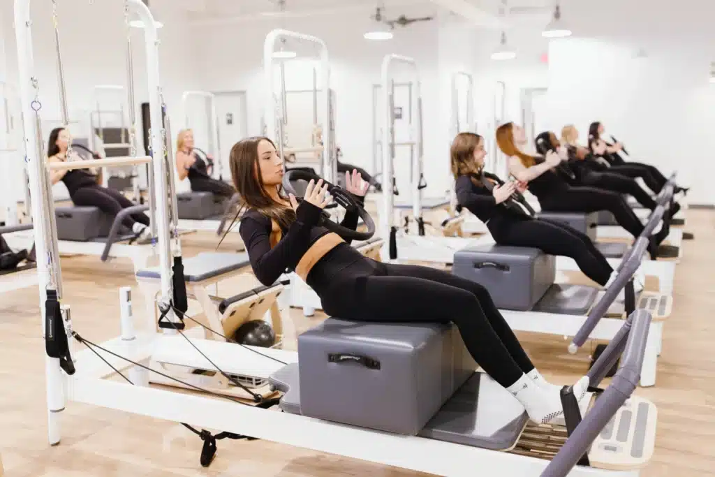 Women in black workout clothes use reformer Pilates machines in a bright, modern fitness studio with wood flooring.