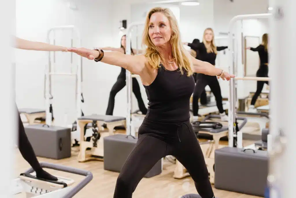 Women in black workout clothes do a yoga pose in a bright, modern fitness studio with exercise equipment.