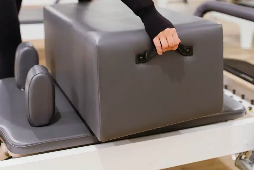 Close-up of a person lifting a gray padded box with a handle, used for Pilates exercise equipment.