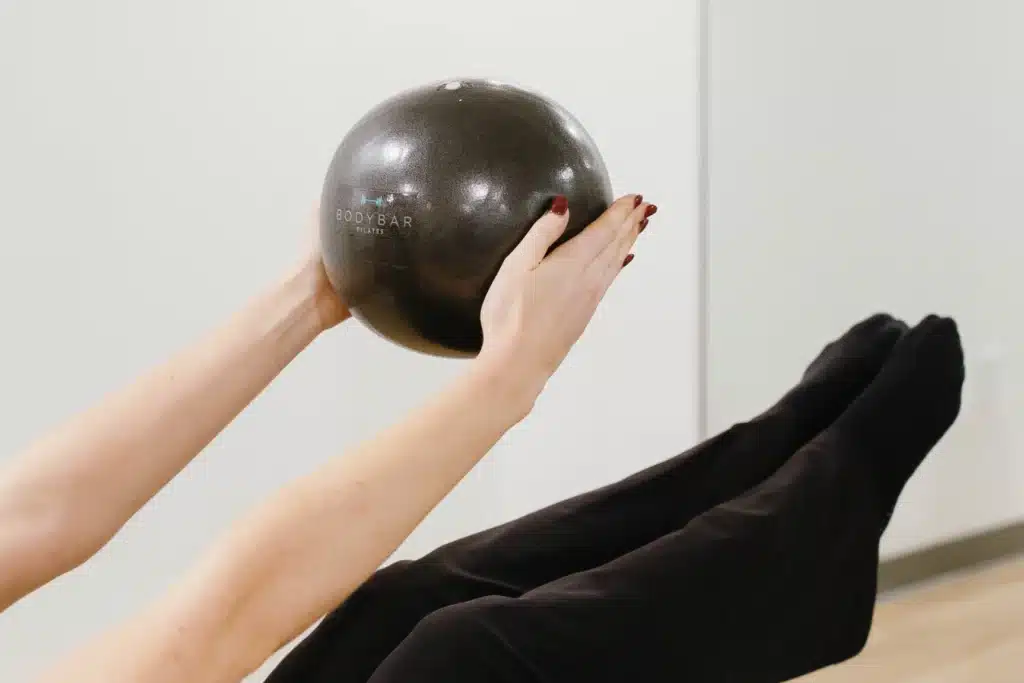 Person holding a black exercise ball while lifting legs, wearing black leggings, in a fitness studio.