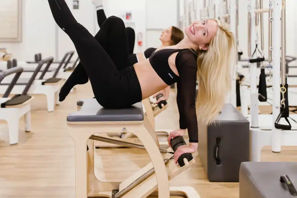 Woman in black workout clothes smiles while exercising on Pilates equipment in a bright studio.