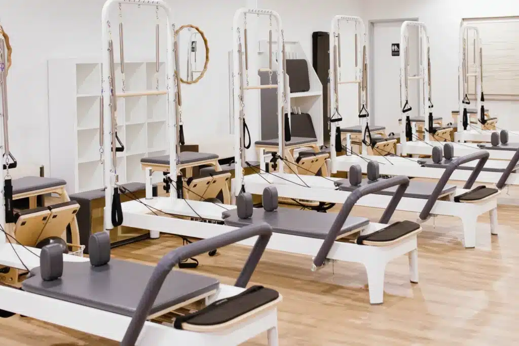 A bright fitness studio with several Pilates reformer machines lined up on a wooden floor.