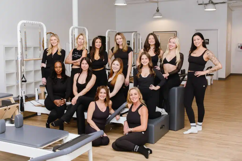 Thirteen women in black athletic wear pose smiling in a bright, modern pilates studio with equipment.
