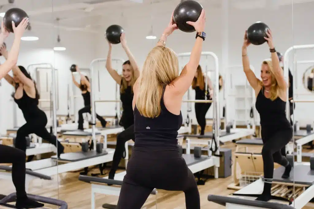 Woman in workout clothes lifts a ball in a fitness studio with mirrors and exercise machines.