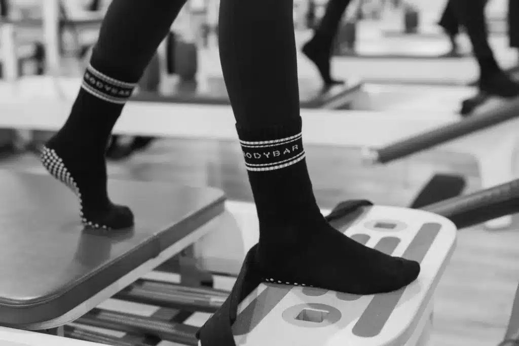 Person wearing grip socks standing on a Pilates reformer machine in a fitness studio.