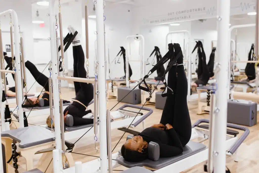 People in black workout attire using Pilates reformer machines in a bright fitness studio, legs raised in straps.