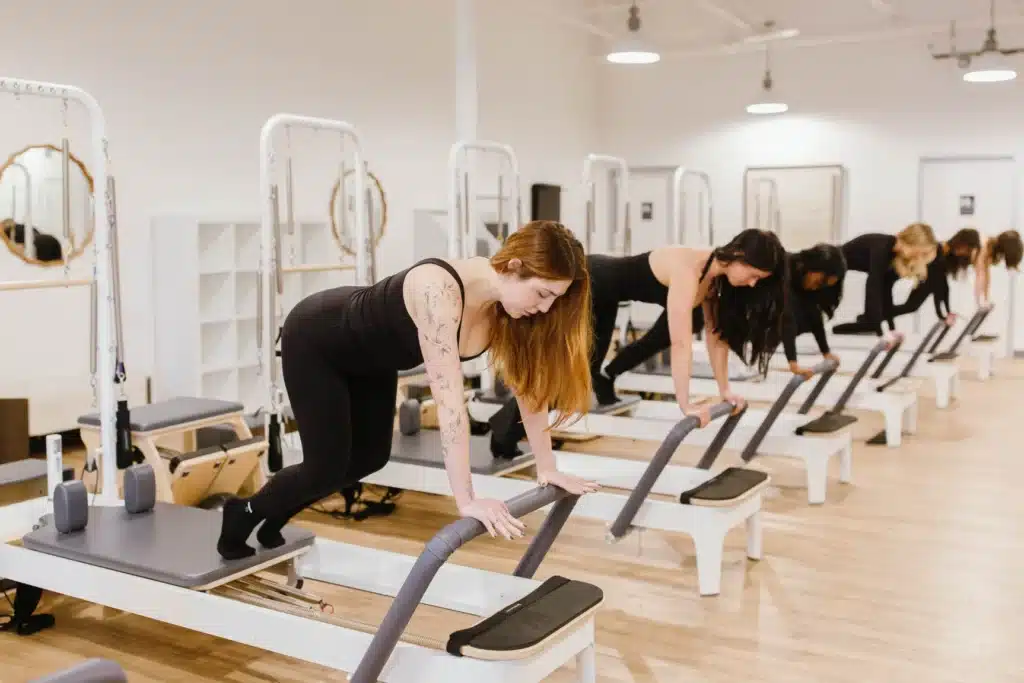 Five women in black workout clothes doing Pilates exercises on reformer machines in a bright studio.
