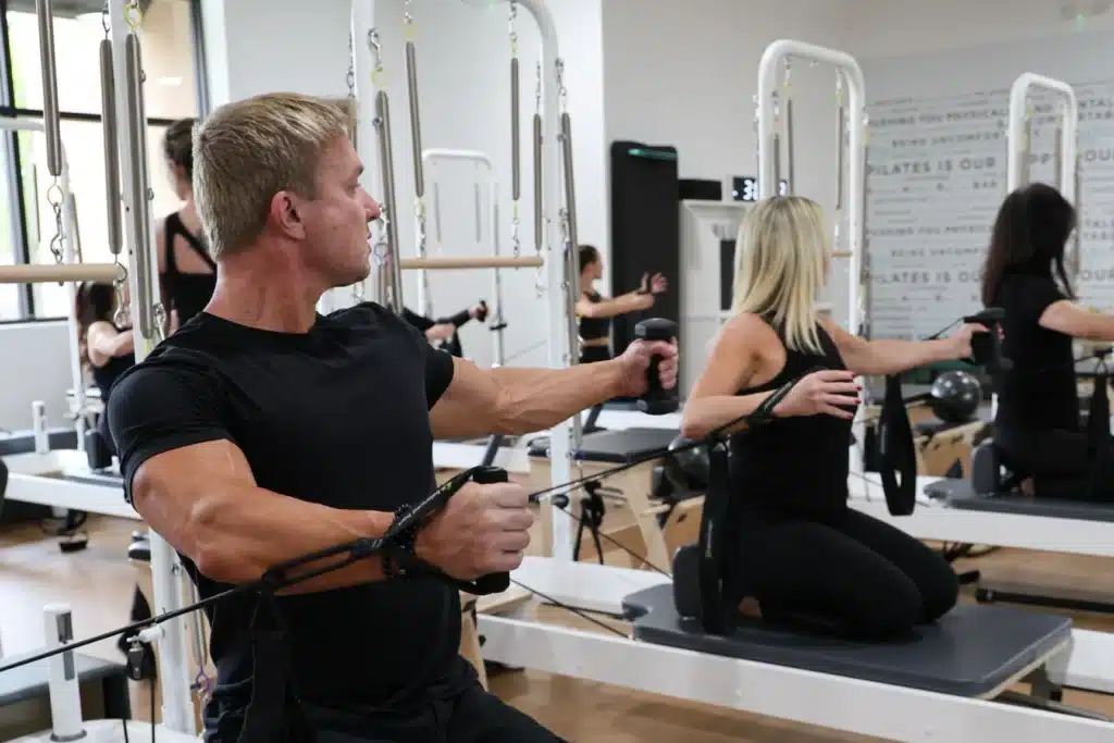 People exercising on Pilates reformer machines in a fitness studio, wearing black workout clothes.