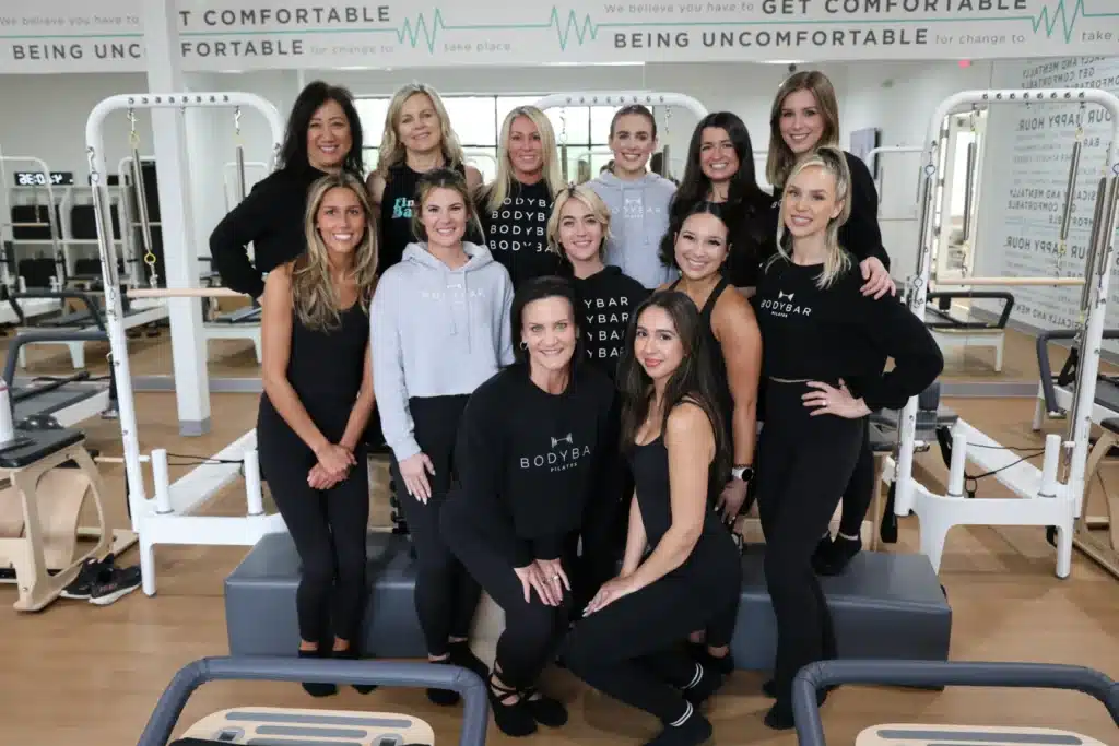 Thirteen women in workout attire smile in a bright fitness studio with Pilates equipment in the background.