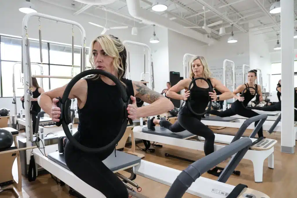Three women in black workout clothes use reformer machines in a bright Pilates studio, each holding a Pilates ring.
