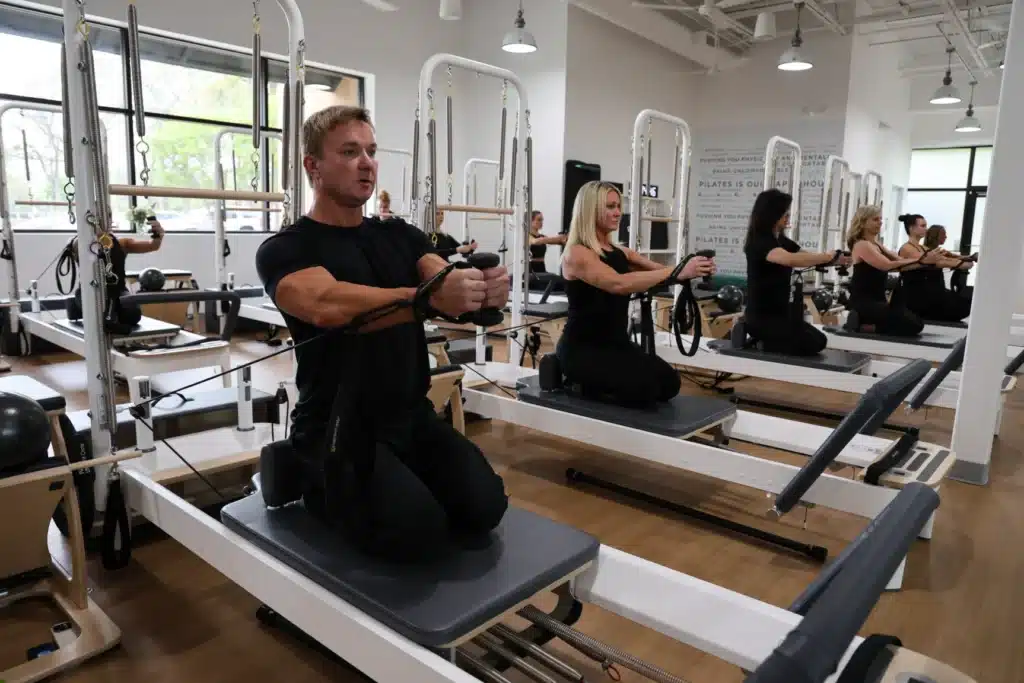People kneeling on Pilates reformers in a bright studio, exercising with resistance bands.
