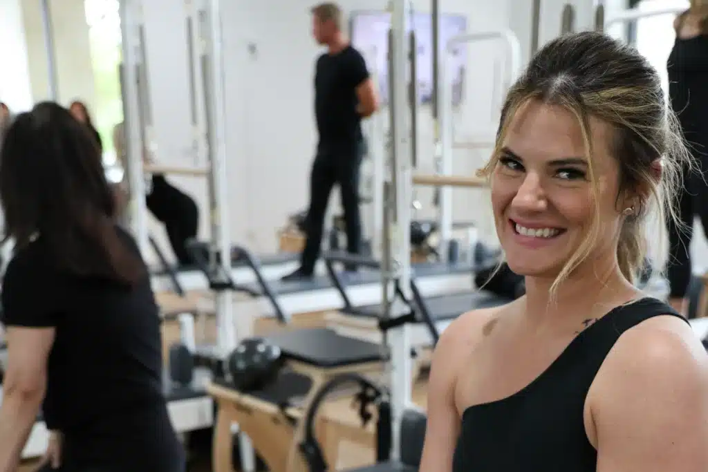 Woman in black workout clothes smiles in a gym, with people exercising on equipment in the background.