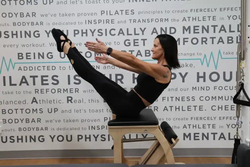 Woman in black workout clothes balances in a Pilates pose on a chair, against a fitness-themed wall background.