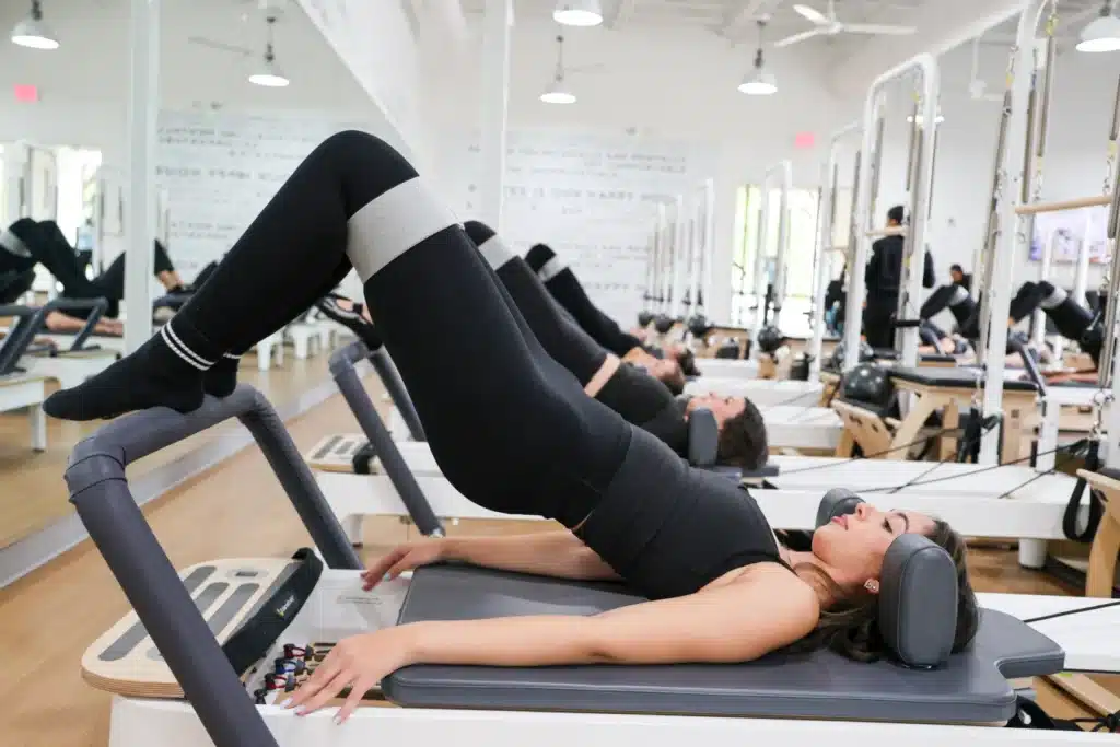 Women in workout clothes do Pilates bridge exercises on reformer machines in a bright fitness studio.