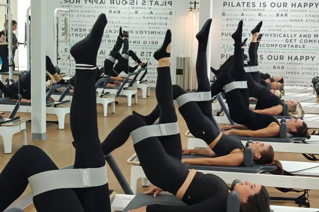 People in black workout clothes do Pilates on reformer machines with resistance bands, raising their legs in a mirrored studio.