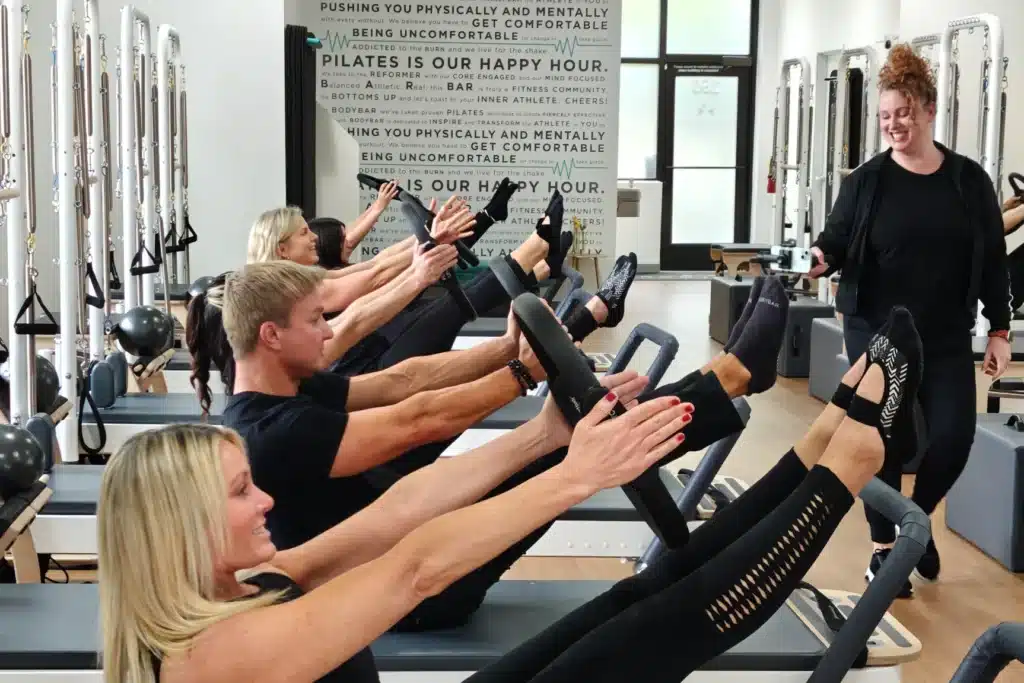 Four people do Pilates on reformer machines as an instructor watches and smiles in a bright studio.