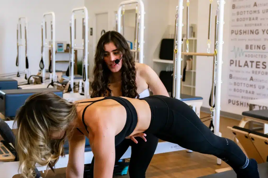 Instructor with headset assists woman in workout at a Pilates studio with reformer machines in the background.