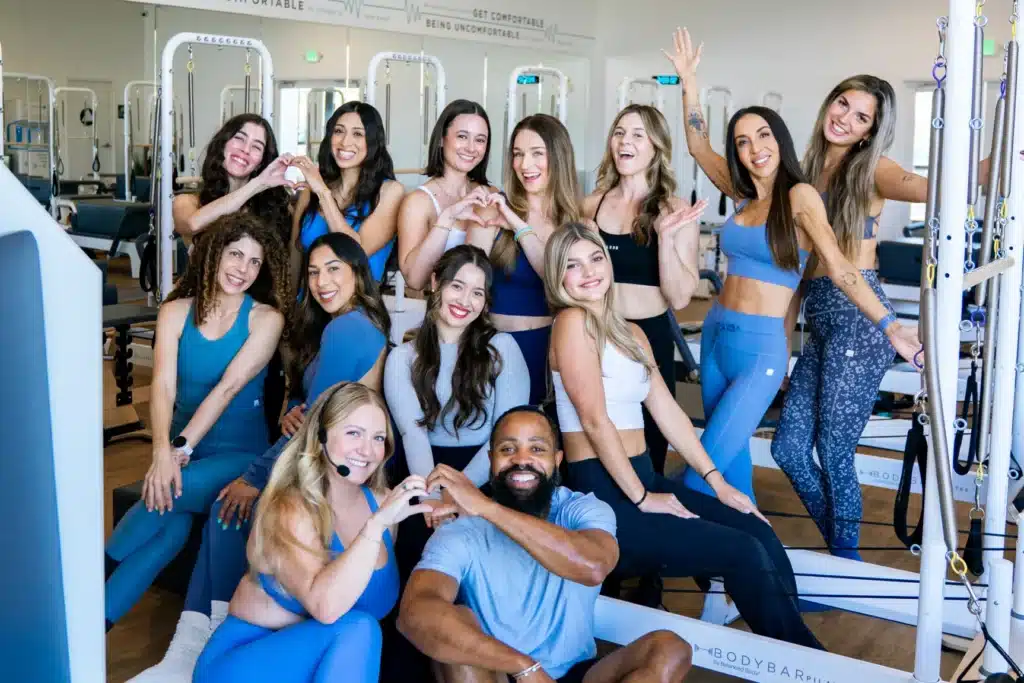 A group of people in athletic wear pose together, smiling and making heart shapes with their hands in a gym.