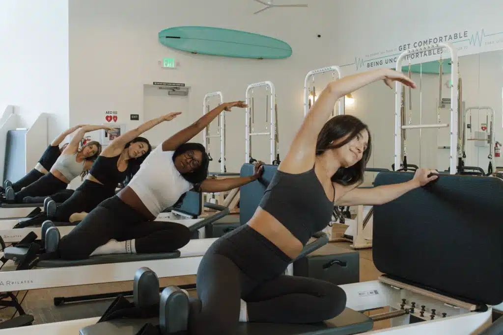 Four women in workout clothes stretch sideways on Pilates reformer machines in a bright fitness studio.