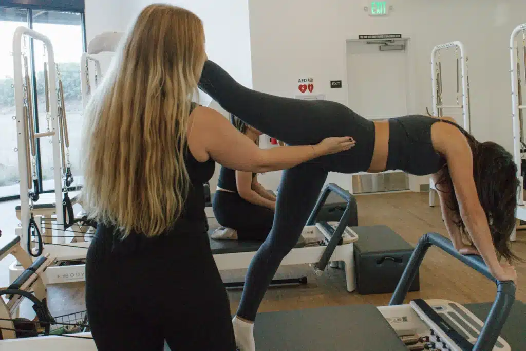 A woman assists another woman balancing on a Pilates reformer in a fitness studio.