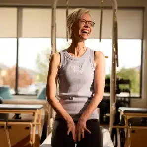 Woman with glasses smiling and sitting on Pilates equipment in a sunlit studio.