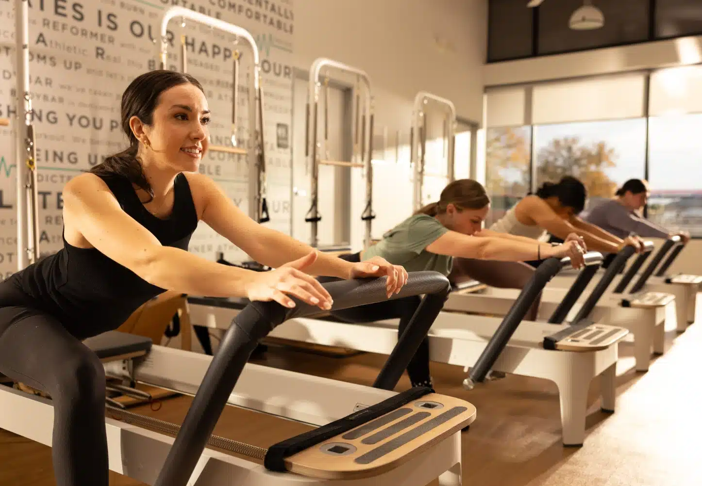 Four people exercise on reformer Pilates machines in a bright studio, with focus on a woman in front wearing black.