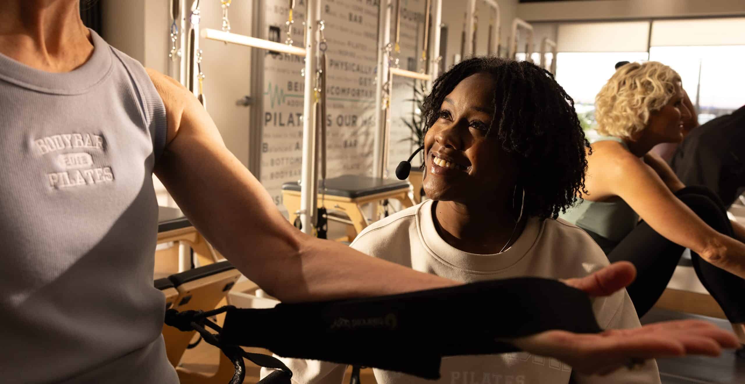 Instructor with headset smiles at a person stretching their arm in a well-lit pilates studio.
