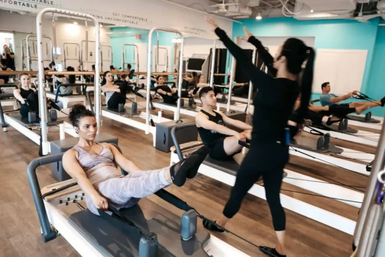 People doing pilates on reformer machines in a bright studio, with an instructor guiding the class.