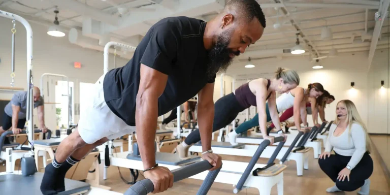 A group of people in a bright studio doing Pilates exercises on reformer machines, led by an instructor.