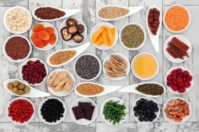 Assorted bowls of seeds, nuts, dried fruits, berries, spices, and chocolate on a rustic white wooden surface.