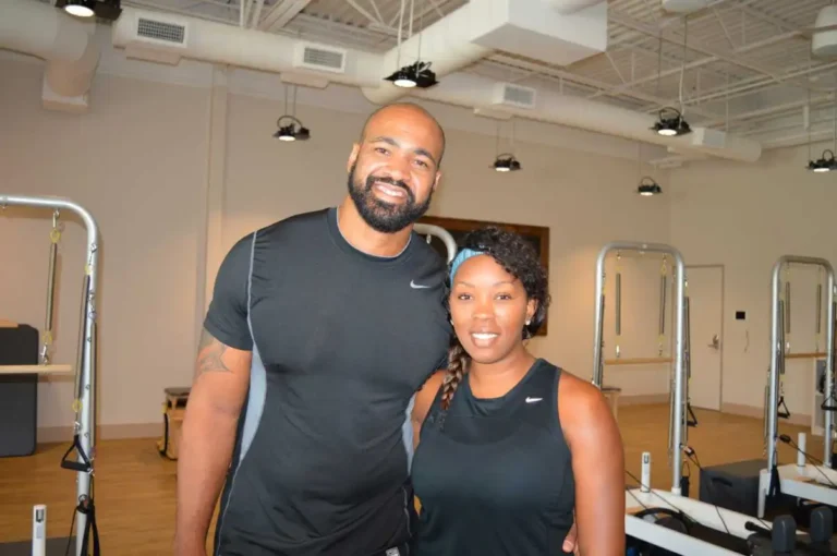 Two people in black athletic wear smile in a gym with Pilates equipment and light-colored walls behind them.