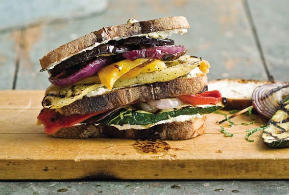 A stacked sandwich with grilled vegetables and cheese on rustic bread, placed on a wooden board.