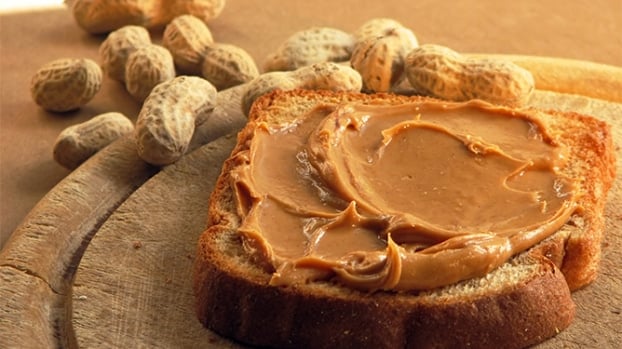 A slice of bread with creamy peanut butter on a wooden board, with unshelled peanuts in the background.