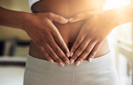 A person forms a heart shape with their hands over their bare stomach, wearing light-colored pants.