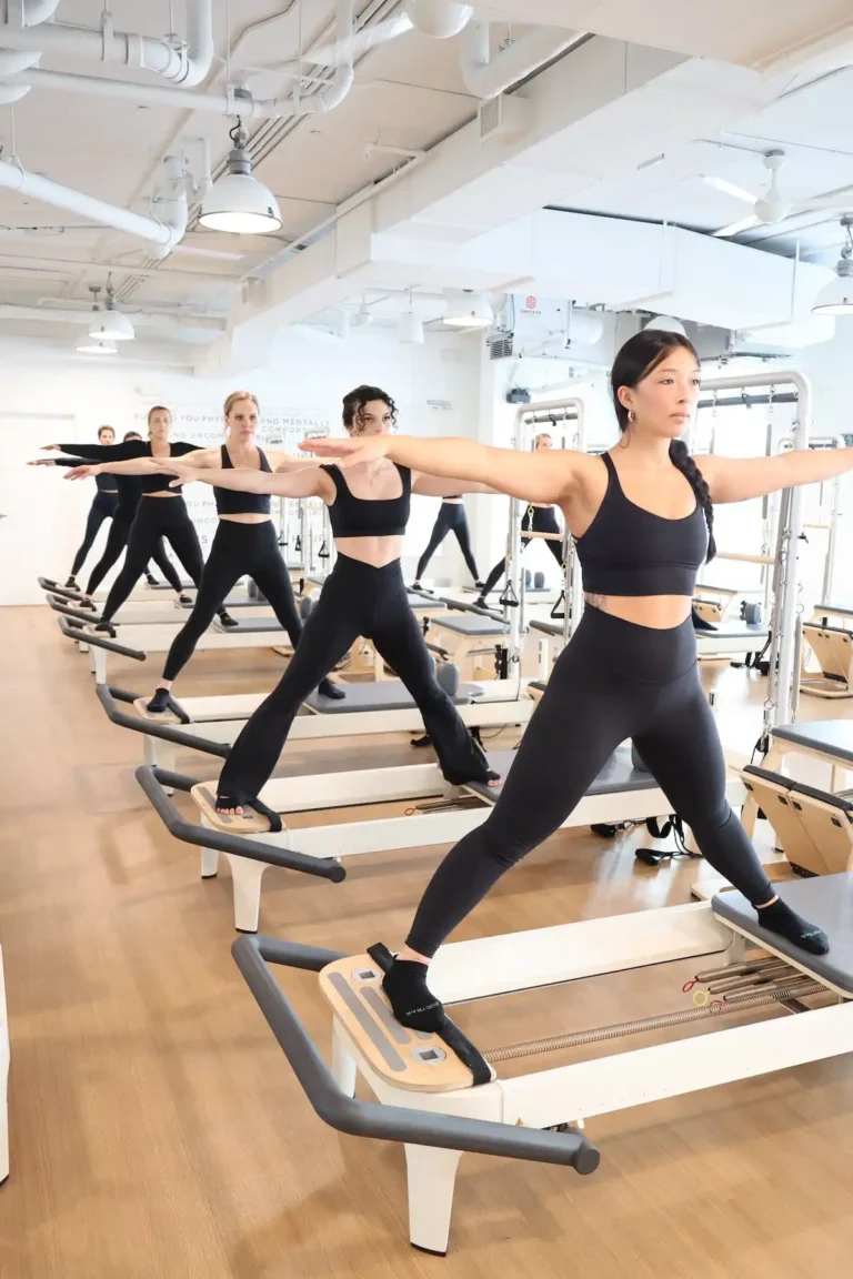 Five women doing Pilates on reformer machines in a bright studio, all wearing black workout clothes, arms extended.