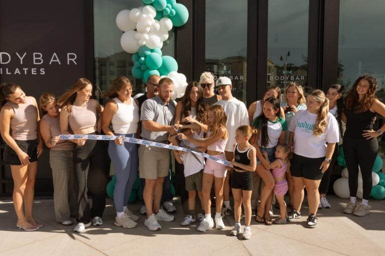 A group of people gathers outside a building, smiling and cutting a ribbon for a grand opening event.