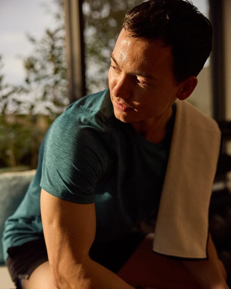 Man in a teal shirt with a white towel on his shoulder sits and sweats, looking to the side in natural light.