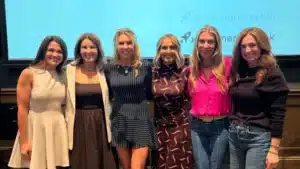 Six women stand together smiling in front of a screen displaying partially visible text and graphics.