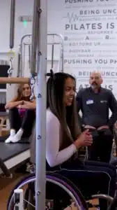 Woman in a wheelchair leads a fitness class, smiling, with two other people in the background at a gym.