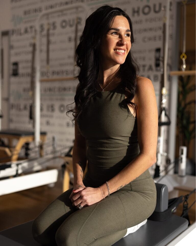 Woman in olive workout clothes sits on exercise equipment, smiling, with sunlight on her face in a fitness studio.