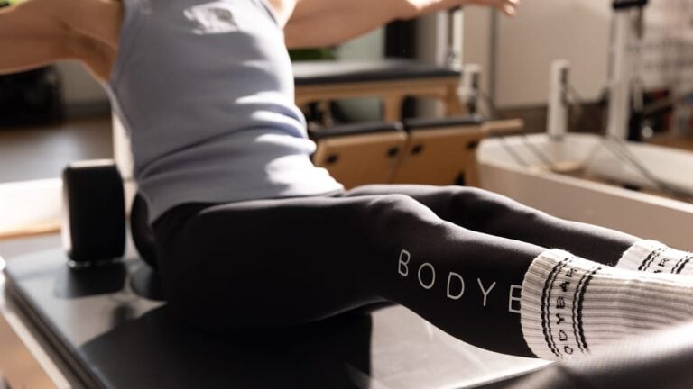 Person in athletic wear exercises on Pilates equipment, legs extended, wearing Body brand leggings and white socks.
